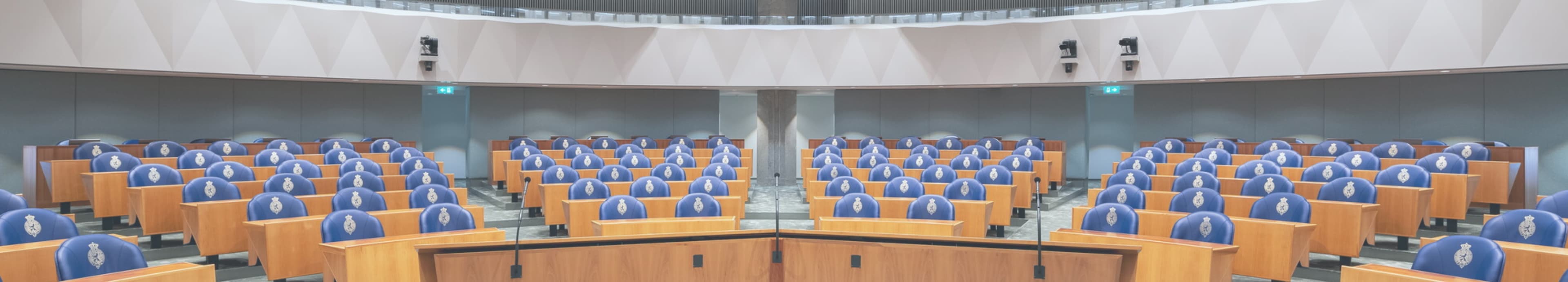 De lege plenaire zaal van de Tweede Kamer: rijen blauwe stoelen met het rijkswapen achter houten bankjes met microfoons.